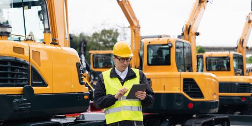 Engineer in a helmet with a digital tablet stands next to construction excavators